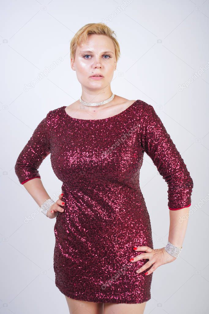 cute caucasian curvy girl with short blonde hair and plus size body wearing beautiful elegant cherry color dress with sequins and posing on a white background in the Studio alone. woman in party cloth