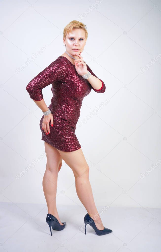 cute caucasian curvy girl with short blonde hair and plus size body wearing beautiful elegant cherry color dress with sequins and posing on a white background in the Studio alone. woman in party cloth