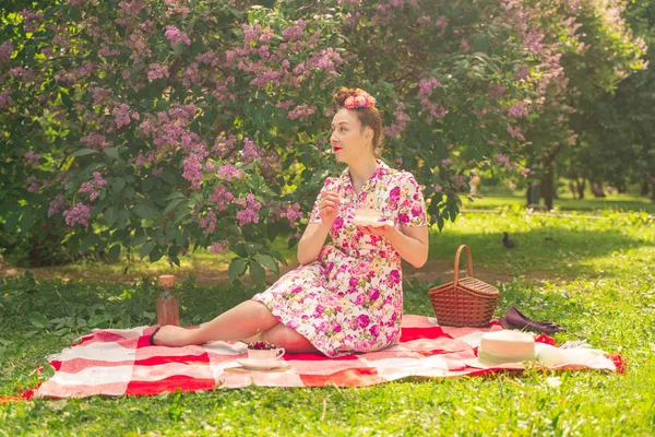 ライラックの茂みの近くの公園でチェッカー毛布に夏のドレスで恋人魅力的なピンナップの女の子は 一人で生活とレジャーを楽しんでいます 草の上にかなり幸せな白人女性 — ストック写真