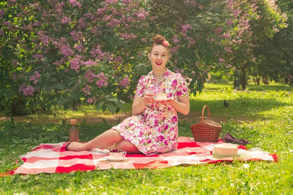 ライラックの茂みの近くの公園でチェッカー毛布に夏のドレスで恋人魅力的なピンナップの女の子は 一人で生活とレジャーを楽しんでいます 草の上にかなり幸せな白人女性 — ストック写真