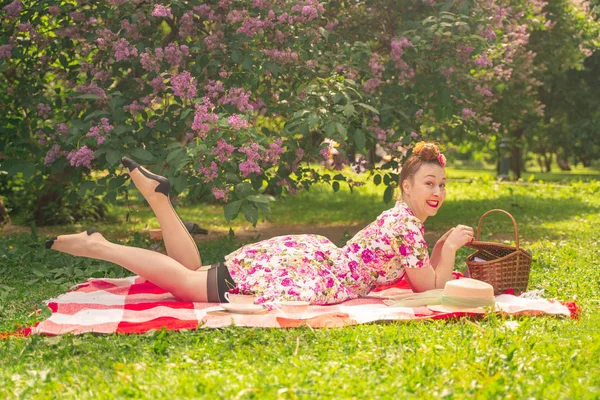 ライラックの茂みの近くの公園でチェッカー毛布に夏のドレスで恋人魅力的なピンナップの女の子は 一人で生活とレジャーを楽しんでいます 草の上にかなり幸せな白人女性 — ストック写真