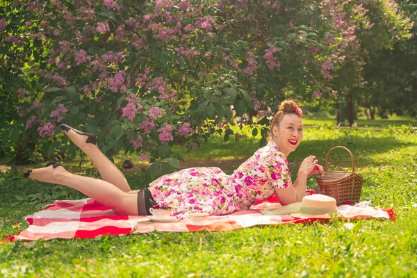 ライラックの茂みの近くの公園でチェッカー毛布に夏のドレスで恋人魅力的なピンナップの女の子は 一人で生活とレジャーを楽しんでいます 草の上にかなり幸せな白人女性 — ストック写真