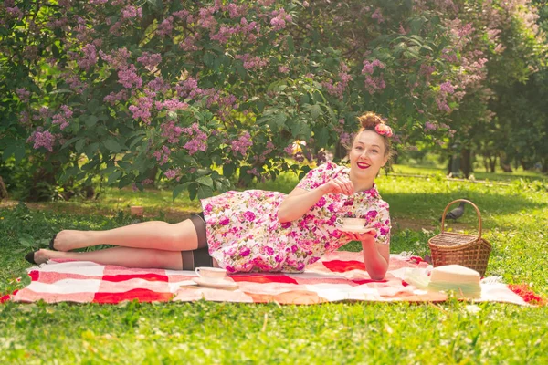 ライラックの茂みの近くの公園でチェッカー毛布に夏のドレスで恋人魅力的なピンナップの女の子は 一人で生活とレジャーを楽しんでいます 草の上にかなり幸せな白人女性 — ストック写真