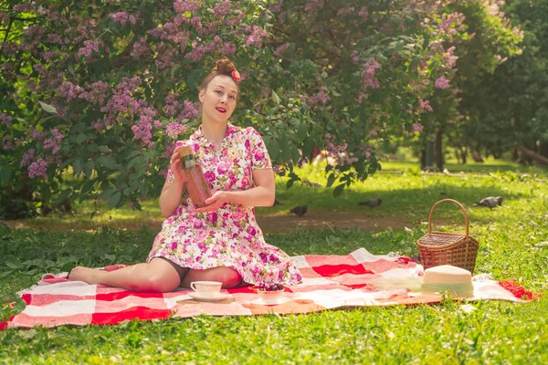 ライラックの茂みの近くの公園でチェッカー毛布に夏のドレスで恋人魅力的なピンナップの女の子は 一人で生活とレジャーを楽しんでいます 草の上にかなり幸せな白人女性 — ストック写真
