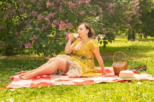 可爱加大小白种女孩穿着一个可爱的复古黄色礼服 享受在绿草的夏季公园度假 弯曲的针脚妇女复古礼服有休息附近的丁香自然 — 图库照片