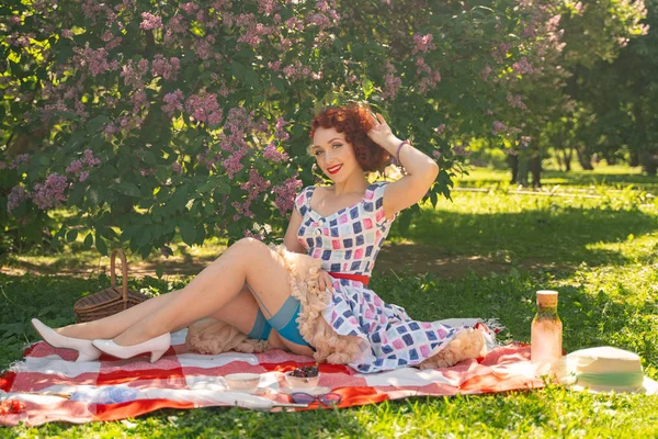 ヴィンテージの夏のドレスと背中に縫い目を持つ古典的なストッキングの赤毛の幸せなピンアップの女の子は ライラックブッシュの隣に 市内の公園の緑の草の上に毛布の上に座っています — ストック写真