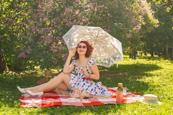 ヴィンテージの夏のドレスと背中に縫い目を持つ古典的なストッキングの赤毛のかわいいピンナップの女性は ライラックブッシュの隣の都市公園の草の上に毛布の上に座っています 太陽の傘を持つ幸せな女の子 — ストック写真