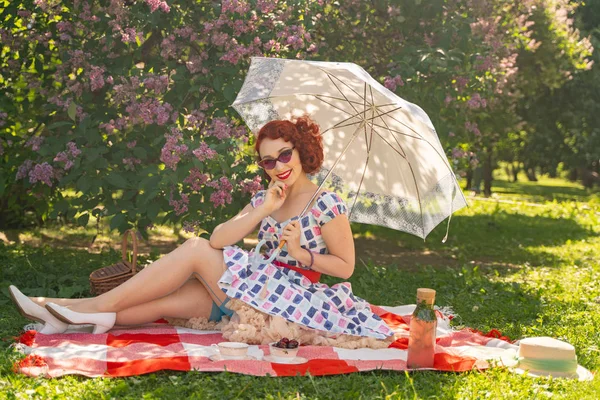 红发漂亮的小号女人穿着复古的夏装和经典的丝袜 后面有一个接缝 坐在城市公园的草地上的毯子上 旁边是丁香布什 快乐的女孩与太阳伞 — 图库照片