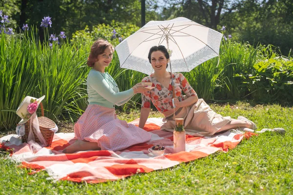 一緒に晴れた日に市立公園で素敵なピクニックを持っている2かわいいピンアップ女性 女の子の友人は暑い夏の天気を楽しみます レトロなヴィンテージスタイルの美しい女性は 草の上でリラックスしています — ストック写真