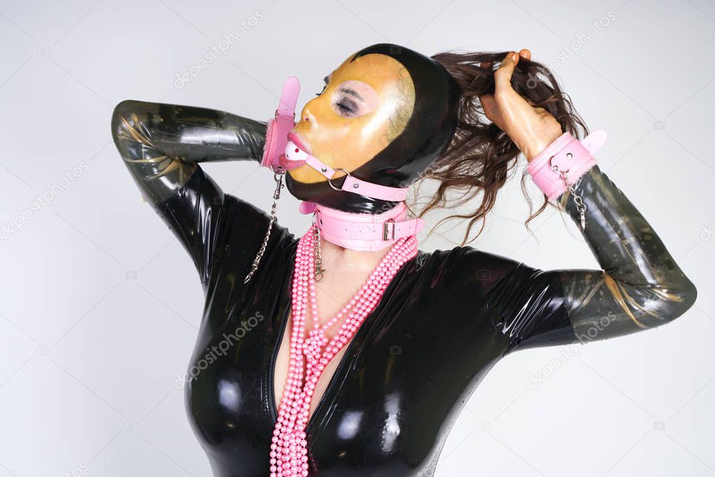 fashionable adult person wearing latex mask with transparent face, rubber catsuit, bdsm pink handcuffs and collar. kinky stylish woman playing with ball gag alone on white studio background.