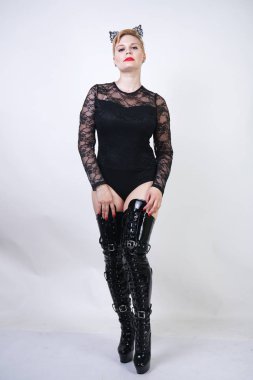 pretty plus size young female with short blonde hair posing on white studio background. hot chubby fashion girl in black lace bodysuit and patent leather thigh high boots standing as cat woman alone. clipart