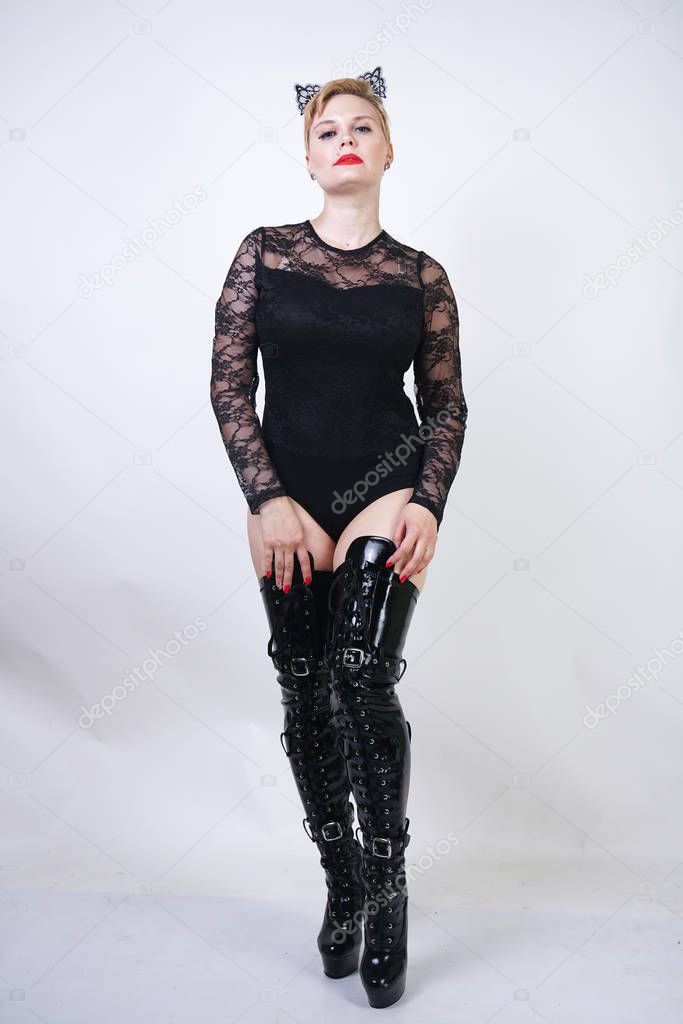 pretty plus size young female with short blonde hair posing on white studio background. hot chubby fashion girl in black lace bodysuit and patent leather thigh high boots standing as cat woman alone.