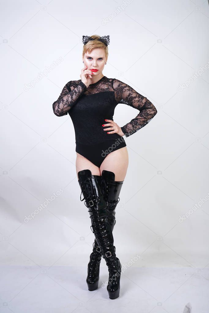 pretty plus size young female with short blonde hair posing on white studio background. hot chubby fashion girl in black lace bodysuit and patent leather thigh high boots standing as cat woman alone.