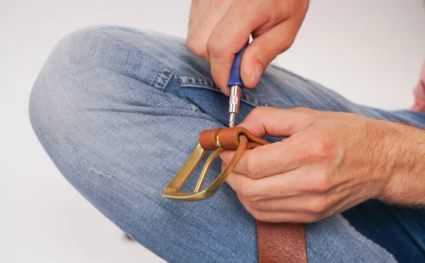 man makes by hands leather belt with buckle. handmade hobby. young man resting by manufacture his designer belts. man with a screwdriver in his hands twists rivets close up on white studio background.