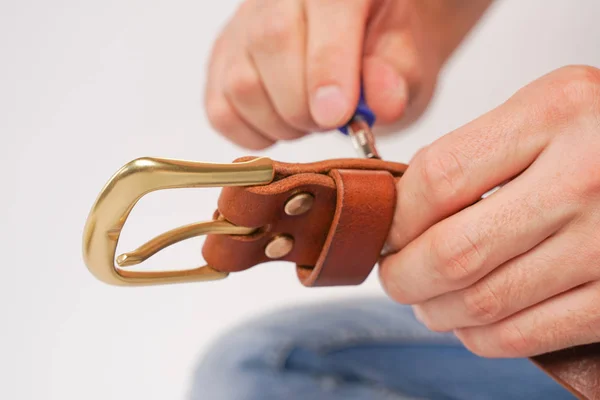 man makes by hands leather belt with buckle. handmade hobby. young man resting by manufacture his designer belts. man with a screwdriver in his hands twists rivets close up on white studio background.