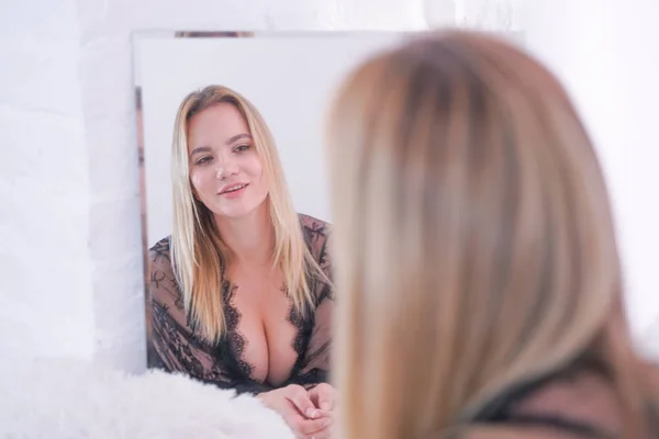 Charmant plus size Kaukasische blonde vrouw gekleed in mooie kant zwarte lingerie en kijken naar zichzelf in de spiegel thuis alleen — Stockfoto