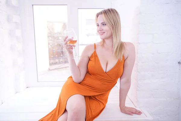 beautiful girl with plus size figure sitting in an orange dress on the windowsill at home and drinking alcohol white wine from a large glass