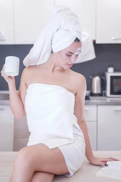 Young girl sitting in the kitchen after a shower wrapped in a white towel and resting — Stockfoto