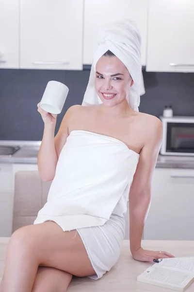 Young girl sitting in the kitchen after a shower wrapped in a white towel and resting — Stockfoto