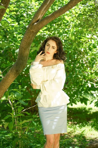 Όμορφη νεαρή μελαχρινή κορίτσι σε λευκό φόρεμα κρύψει από τον καυτό ήλιο στη σκιά του δέντρου στο πάρκο της πόλης — Φωτογραφία Αρχείου