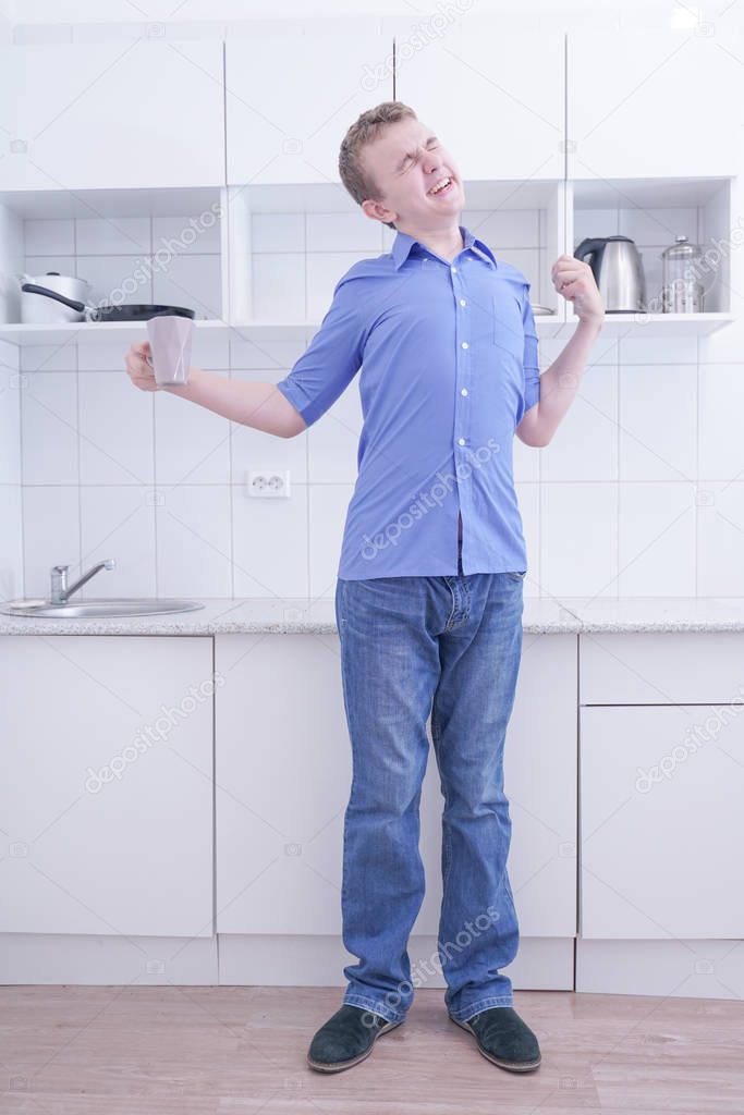 Young bored teenager very tired and stand with cup of coffee at his white kitchen