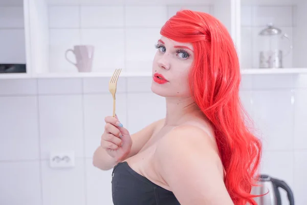 Girl in black corset with red hair holding fork in white kitchen — Stock Photo, Image