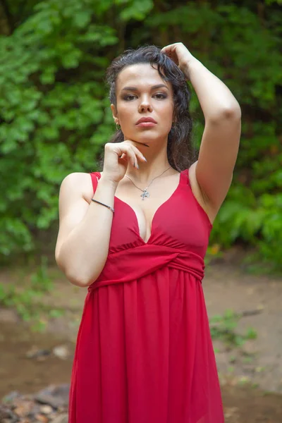 요정 숲에서 포즈를 취하는 세련된 빨간 드레스를 입은 아름다운 소녀. — 스톡 사진