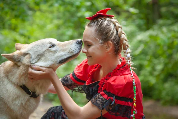 Χαρούμενη όμορφη νεαρή γυναίκα με κόκκινο φόρεμα κάθεται και αγκαλιάζει το λύκο σκυλί της στο δάσος — Φωτογραφία Αρχείου