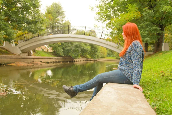 Vacker ung rödhårig kvinna i tröja promenader nära floden i stadsparken — Stockfoto