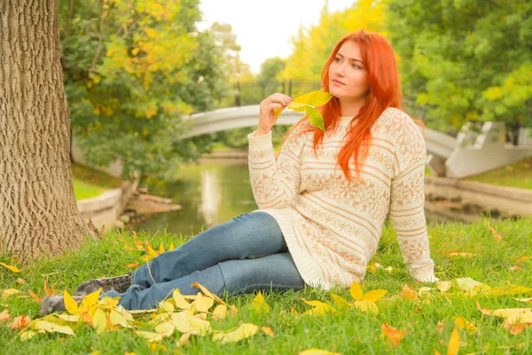 Mulher ruiva em um suéter branco quente do outono sentado no chão perto de uma árvore em um parque da cidade — Fotografia de Stock