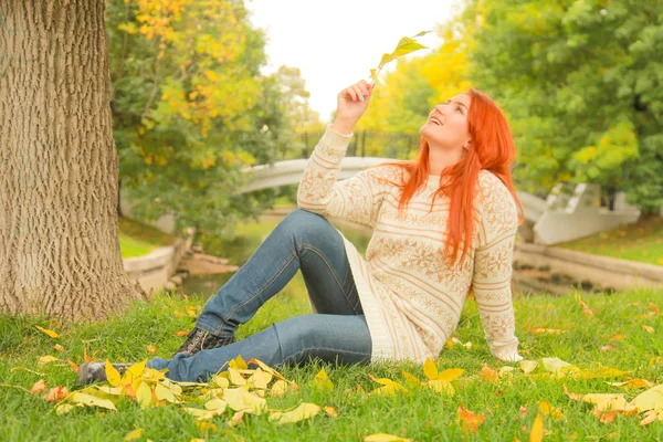 Рыжая женщина в теплом белом осеннем свитере сидит на земле возле дерева в городском парке — стоковое фото