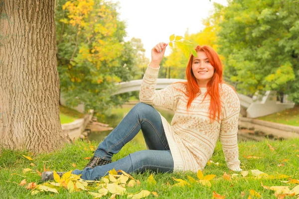 Redhaired woman in a warm white autumn sweater sitting on the ground near a tree in a city Park — Stock Photo, Image