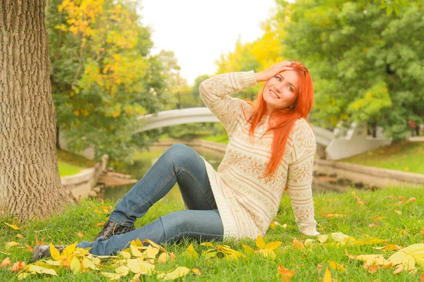 Redhaired nő egy meleg fehér őszi pulóvert ül a földön, közel a fa a városi parkban — Stock Fotó