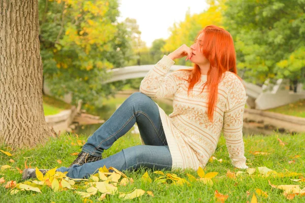 Redhaired nő egy meleg fehér őszi pulóvert ül a földön, közel a fa a városi parkban — Stock Fotó