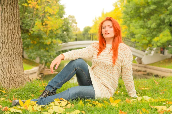 도시 공원의 나무 근처 땅에 앉아 따뜻한 흰색 가을 스웨터에 빨간 머리 여자 — 스톡 사진