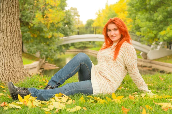 Rothaarige Frau in einem warmen weißen Herbstpullover, die auf dem Boden neben einem Baum in einem Stadtpark sitzt — Stockfoto