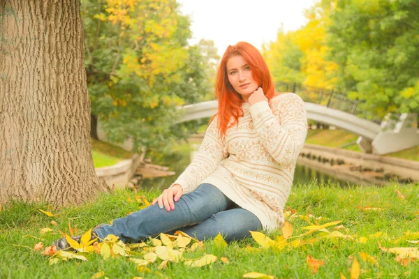 Rothaarige Frau in einem warmen weißen Herbstpullover, die auf dem Boden neben einem Baum in einem Stadtpark sitzt — Stockfoto