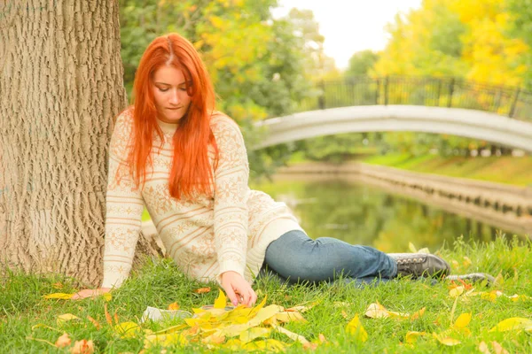 Молодая рыжеволосая девушка читает книгу в городском осеннем зеленом и желтом городском парке — стоковое фото