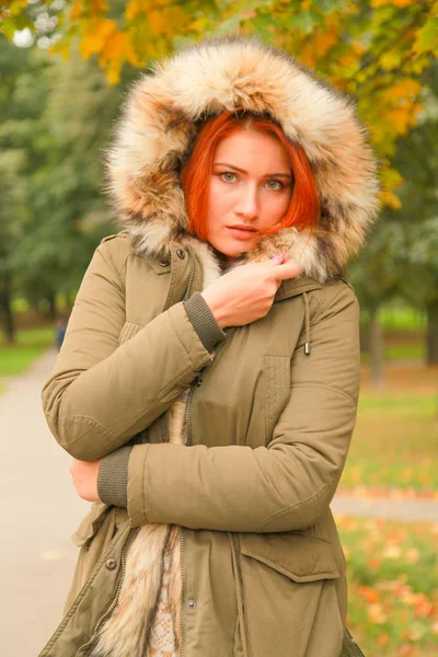 Rothaarige Frau im Mantel mit Pelzhaube im herbstlichen Park mit gelb-grünen Bäumen — Stockfoto