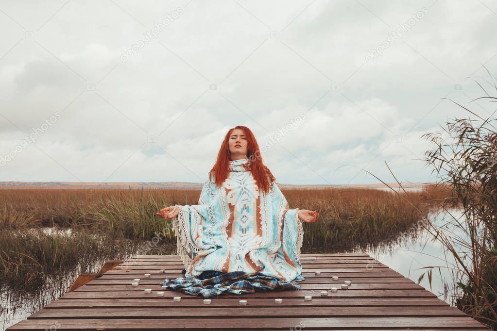 woman practices yoga and meditates with many candles near the autumn lake