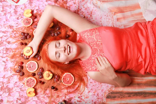 Красива жінка лежить з фруктами навколо волосся на рожевому яскравому фоні — стокове фото