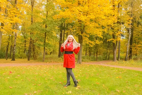 赤いウールのコートで若いかわいいプラスサイズの白人ブロンドの女性は、秋の公園を歩きます — ストック写真