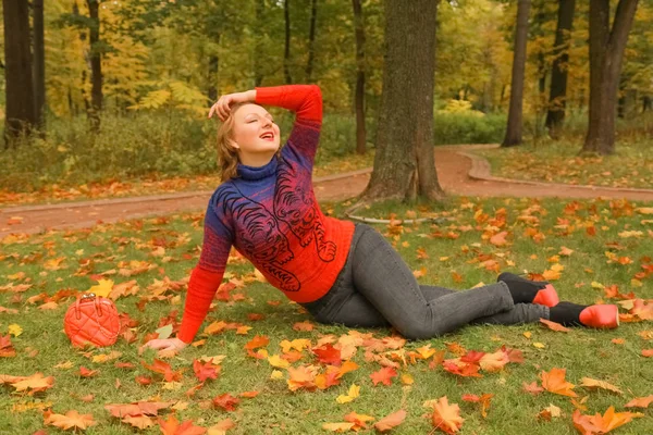 Молодая красивая белая женщина в оранжевом свитере лежит на земле с листьями в осеннем парке — стоковое фото