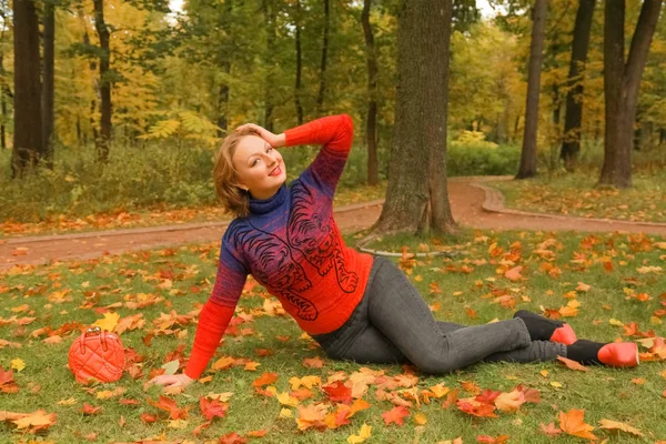 Молодая красивая белая женщина в оранжевом свитере лежит на земле с листьями в осеннем парке — стоковое фото