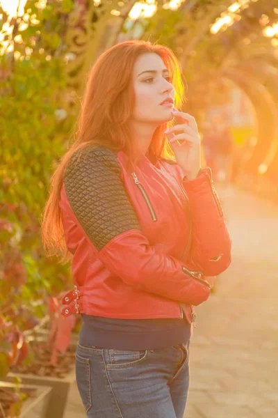 Молодая красивая белая женщина в красной кожаной куртке гуляет в осеннем парке заката — стоковое фото