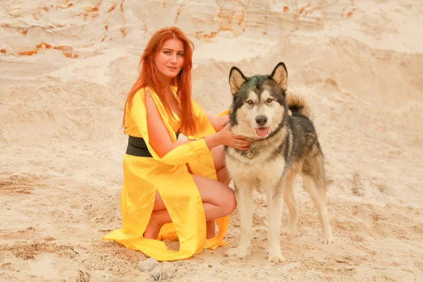 Jovem mulher de cabelo longo posando com seu cão alasca malamute no deserto de areia — Fotografia de Stock