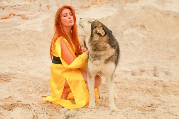 Jovem mulher de cabelo longo posando com seu cão alasca malamute no deserto de areia — Fotografia de Stock