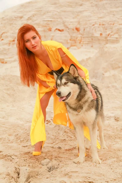 Jovem mulher de cabelo longo posando com seu cão alasca malamute no deserto de areia — Fotografia de Stock