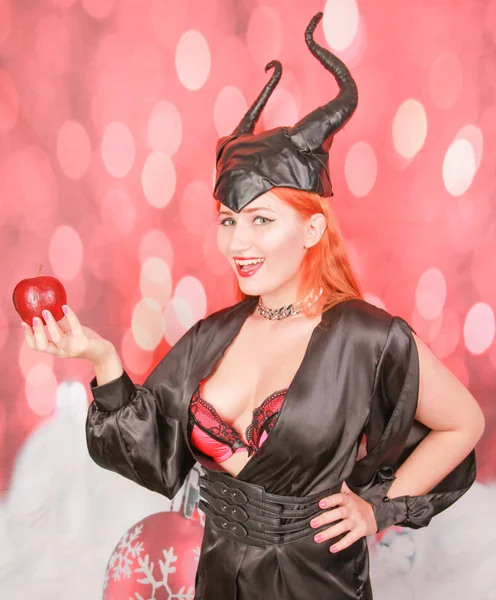 Рыжая девушка в сексуальном черном платье с дьявольскими рогами держит красное яблоко с соблазном и соблазном — стоковое фото