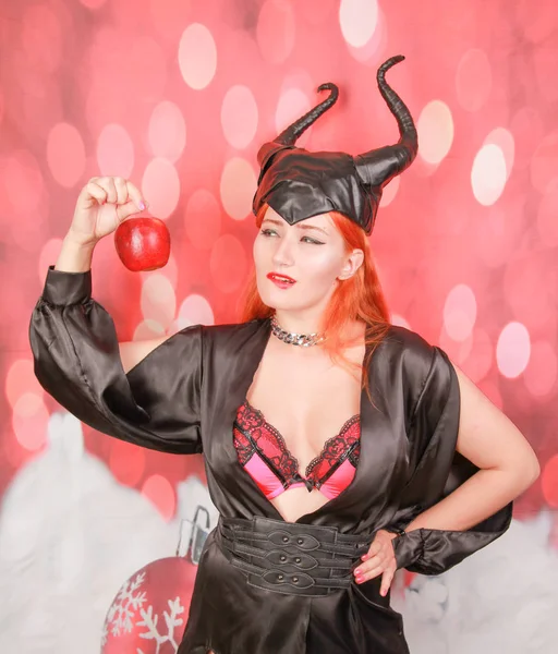 Рыжая девушка в сексуальном черном платье с дьявольскими рогами держит красное яблоко с соблазном и соблазном — стоковое фото